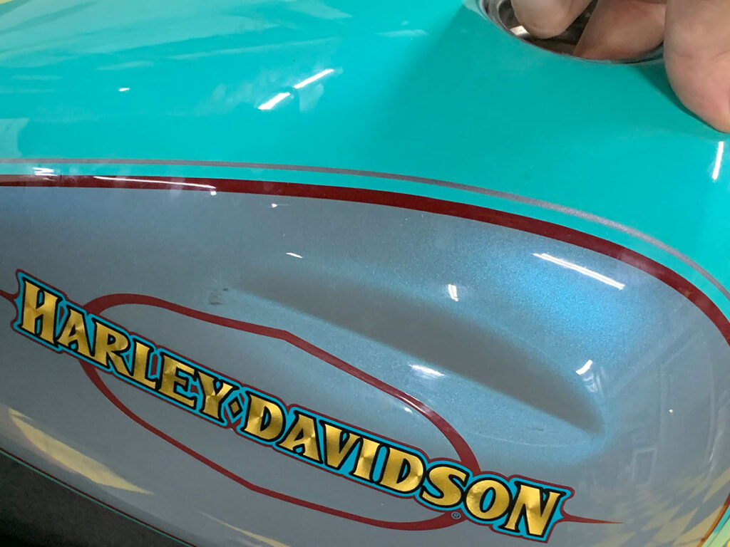 Long, deep dent in a teal and gray Harley Davidson gas tank.