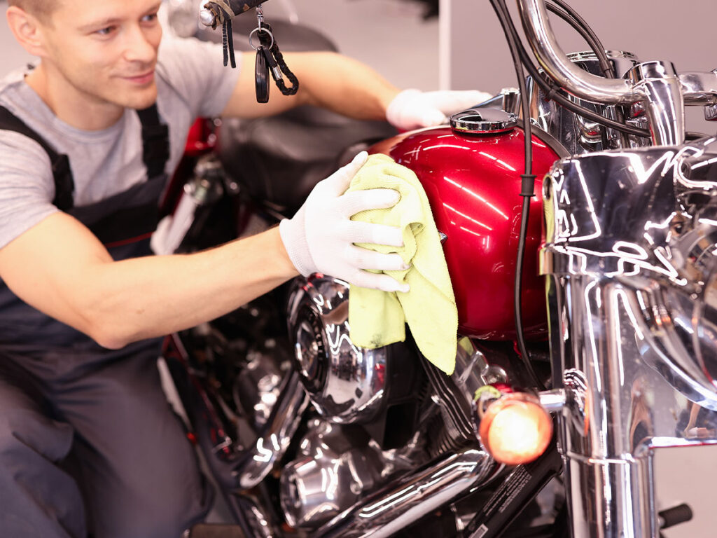 A young man smiling and cleaning the red gas tank of an older motorcycle.